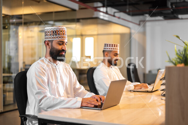التعاون والتشارك في أداء المهام، موظفان عربيان خليجيان عمانيان يرتديان الدشداشة والكمة يجلسان داخل مكتب يقومان بالعمل على الحاسوب المحمول، مهن ووظائف إدارية مكتبية في سلطنة عمان، بيئة عمل مريحة، استخدام الأجهزة التقنية الحديثة في الشركات العمانية