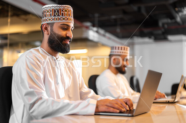 استخدام الأجهزة التقنية الحديثة في الشركات العمانية، التعاون والتشارك في أداء المهام، موظفان عربيان خليجيان عمانيان يرتديان الدشداشة والكمة يجلسان داخل مكتب يقومان بالعمل على الحاسوب المحمول، مهن ووظائف إدارية مكتبية في سلطنة عمان، بيئة عمل مريحة