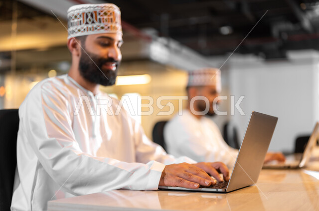 ايماءات البهجة والسرور، استخدام الأجهزة التقنية الحديثة في الشركات العمانية، موظفان عربيان خليجيان عمانيان يرتديان الدشداشة والكمة يجلسان داخل مكتب يقومان بالعمل على الحاسوب المحمول، مهن ووظائف إدارية مكتبية في سلطنة عمان، بيئة عمل مريحة