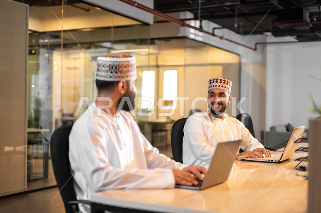 إيماءات البهجة والسرور أثناء تبادل الأحاديث بين الموظفين، استخدام الأجهزة التقنية الحديثة في إنجاز المهام، موظفان عربيان خليجيان عمانيان يرتديان الدشداشة والكمة يجلسان داخل مكتب يقومان باستعمال الحاسوب المحمول، مهن ووظائف إدارية في سلطنة عمان، بيئة عمل مريحة