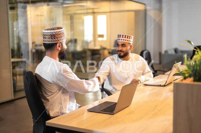 Getting to know colleagues, using modern technology to accomplish tasks, two Gulf Arab Omani employees wearing a dishdasha and a kummah sitting in an office shaking hands with each other, using a laptop in companies, administrative professions and jobs in the Sultanate of Oman, a comfortable work environment