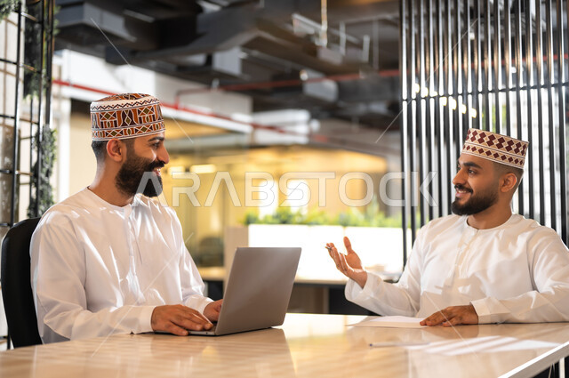 تبادل الأحاديث داخل مقر الشركة، ايماءات البهجة والسرور، استخدام الأجهزة التقنية الحديثة في إنجاز المهام، موظفان عربيان خليجيان عمانيان يرتديان الدشداشة والكمة يجلسان داخل مكتب يقومان باستعمال الحاسوب المحمول، مهن ووظائف إدارية في سلطنة عمان، بيئة عمل مريحة