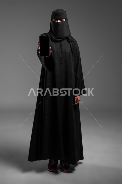 Integrating modern technology into professional life, paying attention to external appearance, looking at the camera with gestures of self-confidence and standing tall, close-up portrait of a veiled Saudi Arabian Gulf Muslim woman wearing the traditional abaya and showing a black blank screen, full-length body, gray background