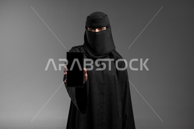 الاهتمام بالمظهر الخارجي والأناقة، النظر للكاميرا بإيمات الثقة بالنفس والوقوف بشموخ، بورتريه مقرب لامرأة عربية خليجية سعودية مسلمة منقبة ترتدي العباءة التقليدية وتعرض شاشة فارغة سوداء، دمج التكنولوجيا الحديثة بالحياة العميلة، خلفية رمادية