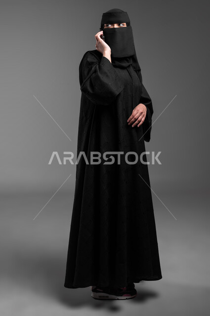 القيام بمكالمة هاتفية صوتية، دمج التكنولوجيا الحديثة بالحياة العميلة، الأناقة والاهتمام بالمظهر الخارجي، بورتريه لامرأة عربية خليجية سعودية مسلمة منقبة ترتدي العباءة التقليدية وتتحدث عبر الجوال، الجسم بالطول الكامل، خلفية رمادية