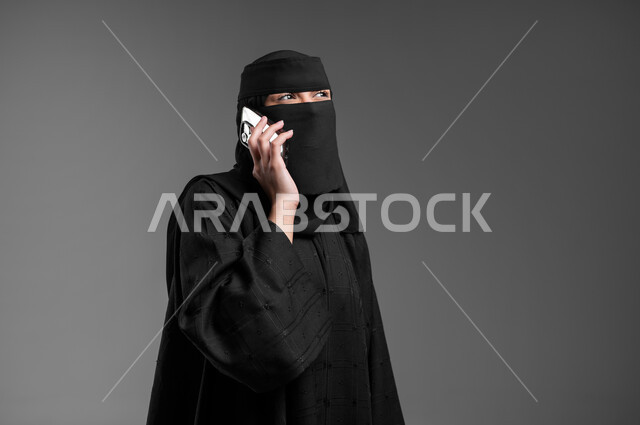 Using a mobile phone to communicate with family and friends, integrating modern technology into customer life, elegance and attention to external appearance, a close-up portrait of a veiled Saudi Arabian Gulf Muslim woman wearing the traditional abaya and making a phone call, gray background