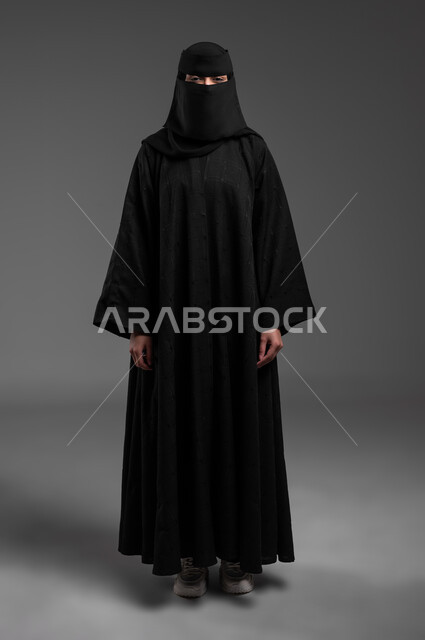 الوقوف بظهر مشدود بإيماءات الثقة بالنفس، مفهوم الحشمة والحياء، الأناقة والاهتمام بالمظهر الخارجي، بورتريه لامرأة عربية خليجية سعودية مسلمة منقبة ترتدي العباءة السوداء التقليدية، صورة الجسم بالطول الكامل، خلفية رمادية