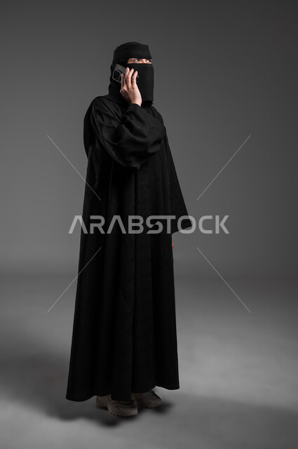 التحدث بالهاتف المحمول والتواصل مع الأهل والأصدقاء، دمج التكنولوجيا الحديثة بالحياة العميلة، الأناقة والاهتمام بالمظهر الخارجي، بورتريه لامرأة عربية خليجية سعودية مسلمة منقبة ترتدي العباءة التقليدية وتجري مكالمة هاتفية، الجسم بالطول الكامل، خلفية رمادية