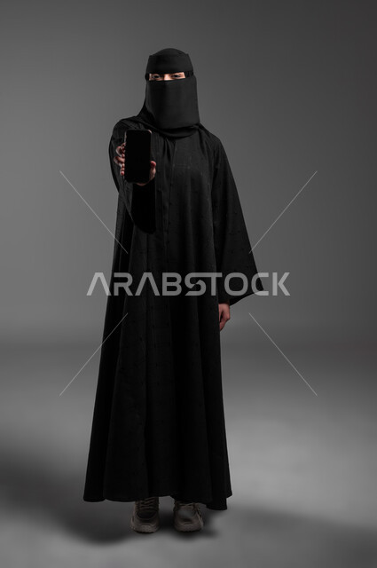 الاهتمام بالمظهر الخارجي، النظر للكاميرا بإيماءات الثقة بالنفس والوقوف بشموخ، بورتريه لامرأة عربية خليجية سعودية مسلمة منقبة ترتدي العباءة التقليدية وتعرض شاشة فارغة سوداء، دمج التكنولوجيا الحديثة بالحياة العميلة، الجسم بالطول الكامل، خلفية رمادية