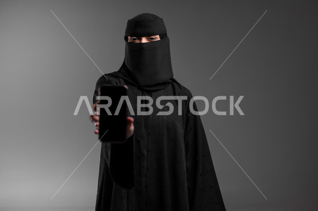 Elegance and attention to external appearance, looking at the camera with gestures of self-confidence and standing proudly, a close-up portrait of a veiled Saudi Gulf Arab Muslim woman wearing the traditional abaya and displaying a blank black screen, integrating modern technology into working life, gray background