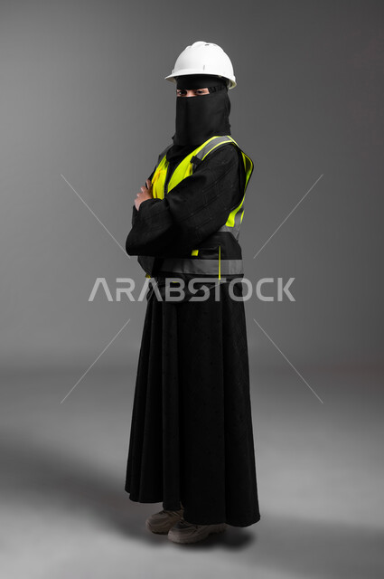 الإعمار بسواعد أبناء الوطن، مهن ووظائف نسائية في القطاع الهندسي الانشائي, بورتريه من الجانب لمهندسة عربية خليجية سعودية مسلمة منقبة ترتدي الخوذة البيضاء وسترة الحماية، الوقوف بظهر مشدود بإيماءات الثقة بالنفس، الجسم بالطول الكامل، خلفية رمادية