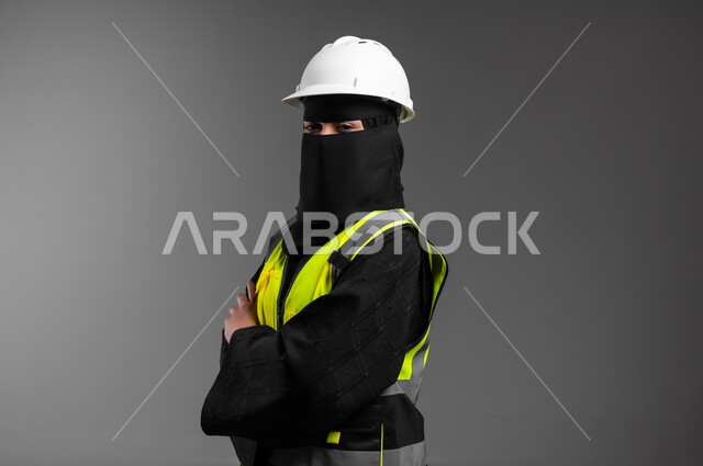 الوقوف بيدين متقاطعتين بإيماءات الثقة بالنفس، دور الهندسة الإنشائية في تطور المجتمع السعودي, مهن ووظائف نسائية في القطاع الهندسي, بورتريه مقرب لمهندسة عربية خليجية سعودية مسلمة منقبة ترتدي الخوذة البيضاء وسترة الحماية، الإعمار بسواعد أبناء الوطن، خلفية رمادية