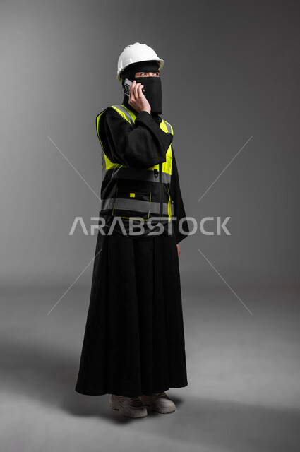 دمج التقنيات التكنولوجية بالقطاع الهندسي، الاستشارة الهاتفية مع فريق العمل، عمل النساء في المجالات الهندسية, بورتريه لمهندسة عربية خليجية سعودية مسلمة منقبة ترتدي الخوذة البيضاء وسترة الحماية تجري مكالمة هاتفية عبر الجوال، الجسم بالطول الكامل، خلفية رمادية