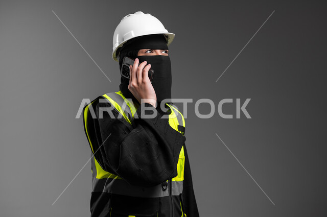الاستشارة الهاتفية والمتابعة مع فريق العمل، دمج التقنيات التكنولوجية بالقطاع الهندسي، عمل النساء في المجالات الهندسية, بورتريه مقرب لمهندسة عربية خليجية سعودية مسلمة منقبة ترتدي الخوذة البيضاء وسترة الحماية تجري مكالمة هاتفية عبر الجوال، خلفية رمادية