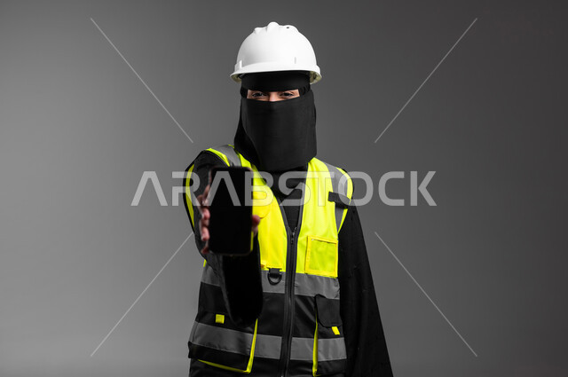 عمل النساء في المجالات الهندسية في السعودية, عرض شاشة جوال سوداء فارغة، دمج التقنيات التكنولوجية بالقطاع الهندسي، بورتريه مقرب لمهندسة عربية خليجية سعودية مسلمة منقبة ترتدي الخوذة البيضاء وسترة الحماية تمسك الهاتف المحمول بيدها، خلفية رمادية