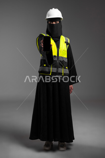Female engineering jobs in the Kingdom, displaying a blank black mobile screen, integrating technological technologies into the engineering field, portrait of a veiled Saudi Gulf Arab Muslim engineer wearing a white helmet and protective jacket holding a mobile phone in her hand, full-length body, gray background