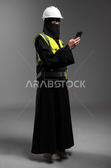 إعمار السعودية بسواعد أبنائها، وظائف نسائية هندسية في المملكة, دمج التقنيات التكنولوجية بالمجال الهندسي، بورتريه لمهندسة عربية خليجية سعودية مسلمة منقبة ترتدي الخوذة البيضاء وسترة الحماية تمسك الهاتف المحمول وتتابع العمل، الجسم بالطول الكامل، خلفية رمادية