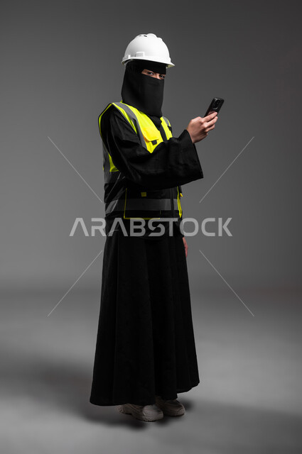 The renaissance of Saudi society with the help of its children, engineering jobs for women in the Kingdom, integration of technological technologies into the engineering field, portrait of a veiled Saudi Gulf Arab Muslim engineer wearing a white helmet and protective vest, holding a mobile phone and continuing to work, full-length body, gray background