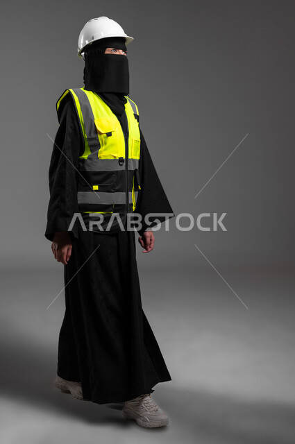 عمل النساء في المجالات الهندسية في المملكة العربية السعودية, بورتريه لمهندسة عربية خليجية سعودية مسلمة منقبة ترتدي الخوذة البيضاء وسترة الحماية تمشي لمتابعة سير العمل والإشراف عليه عن قرب، الجسم بالطول الكامل، خلفية رمادية