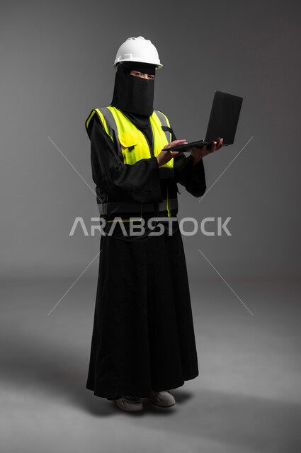مهن ووظائف نسائية هندسية في المملكة, مفهوم إدارة المشاريع وتدقيقها, دمج التكنولوجيا مع المجال الهندسي، بورتريه لمهندسة عربية خليجية سعودية مسلمة منقبة ترتدي الخوذة البيضاء وسترة الحماية تمسك الحاسوب المحمول وتتابع العمل، الجسم بالطول الكامل، خلفية رمادية