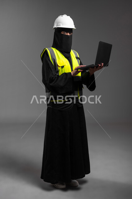 The concept of project management and auditing, integrating technology with the engineering field, female engineering professions and jobs in the Kingdom, portrait of a veiled Saudi Gulf Arab Muslim engineer wearing a white helmet and protective jacket, holding a laptop and continuing work, full-length body, gray background