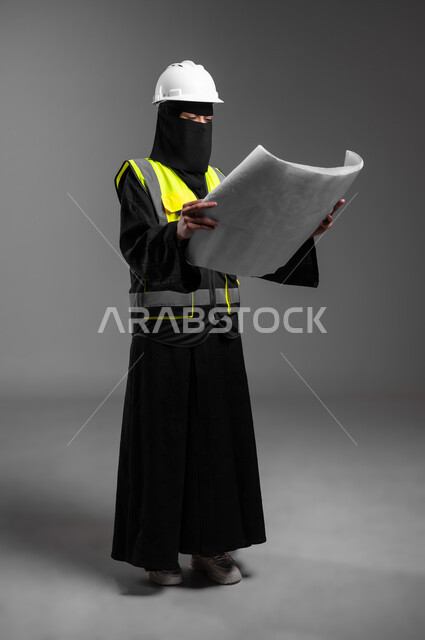 Following up on plans and supervising the progress of work, the role of structural engineering in the development of Saudi society, women’s professions and jobs in the engineering sector, portrait of a veiled Saudi Arabian Gulf Muslim engineer wearing a white helmet and protective vest holding the reconstruction plan, full-length body, gray background