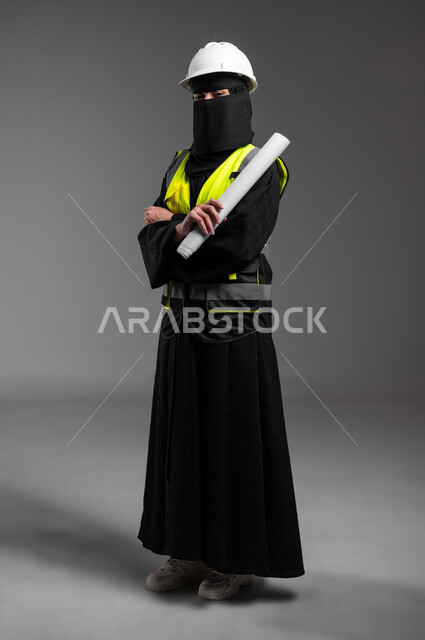 Standing with crossed hands and holding the architectural plan with confidence, the role of structural engineering in the development of Saudi society, women’s professions and jobs in the engineering sector, portrait of a veiled Saudi Gulf Arab Muslim engineer wearing a white helmet and protection vest, full-length body, gray background