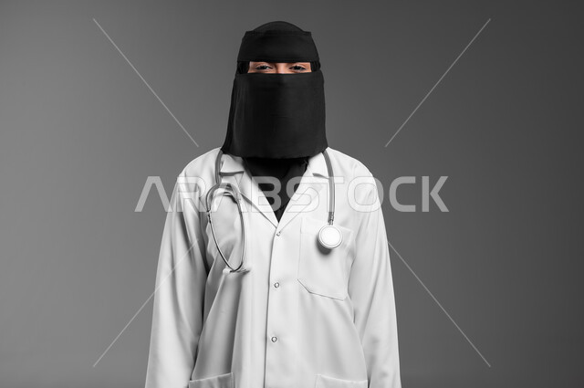 النظر للكاميرا بإيماءات الثقة بالنفس والشموخ، مساهمة النساء في مجال الرعاية الصحية في المملكة العربية السعودية, بورتريه مقرب لطبيبة عربية خليجية سعودية مسلمة منقبة ترتدي المعطف الطبي وتضع سماعة الفحص حول عنقها، خلفية رمادية