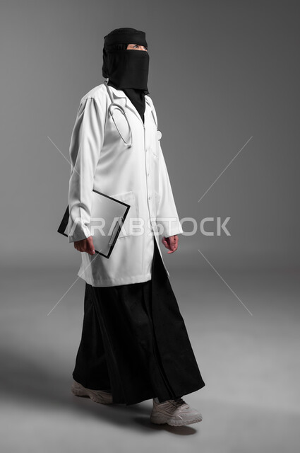 التوجه للإطمئان على صحة المريض، عمل النساء في مجال الرعاية الصحية, كتابة التقارير الطبية، بورتريه لطبيبة عربية خليجية سعودية مسلمة منقبة ترتدي المعطف الطبي وتضع سماعة الفحص حول عنقها تمسك بيدها ملف المرضى، الجسم بالطول الكامل، خلفية رمادية