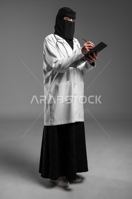 كتابة التقارير الطبية، تدوين وتسجيل الملاحظات حول حالة المريض، عمل النساء في مجال الرعاية الصحية,  بورتريه لطبيبة عربية خليجية سعودية مسلمة منقبة ترتدي المعطف الطبي وتضع سماعة الفحص حول عنقها تمسك بيدها ملف المرضى، الجسم بالطول الكامل، خلفية رمادية