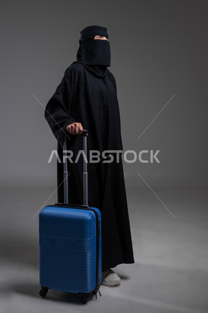 قضاء أوقات ممتعة بالتعرف على بلدان وثقافات أخرى، جر حقيبة السفر والاستعداد للذهاب في الرحلة، مفهوم السياحة في السعودية، بورتريه لامرأة عربية خليجية سعودية مسلمة منقبة ترتدي العباءة السوداء تقف باستقامة، الجسم بالطول الكامل، خلفية رمادية