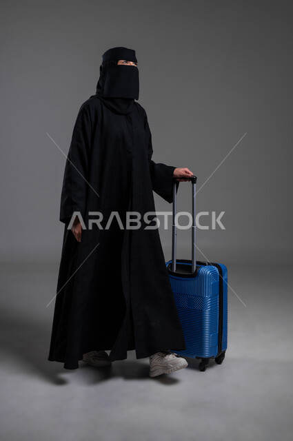 جر حقيبة السفر والاستعداد للذهاب في الرحلة، قضاء أوقات ممتعة بالتعرف على بلدان وثقافات أخرى، مفهوم السياحة في السعودية، بورتريه لامرأة عربية خليجية سعودية مسلمة منقبة ترتدي العباءة السوداء تمشي متجهة للأمام، الجسم بالطول الكامل، خلفية رمادية