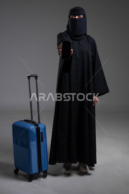 الوقوف بظهر مشدود وعرض شاشة سوداء فارغة، الاستعداد للرحلات السياحية،  بورتريه لامرأة عربية خليجية سعودية مسلمة منقبة ترتدي العباءة السوداء تمسك الهاتف المحمول وبجانبها حقيبة السفر الزرقاء، الجسم بالطول الكامل، خلفية رمادية