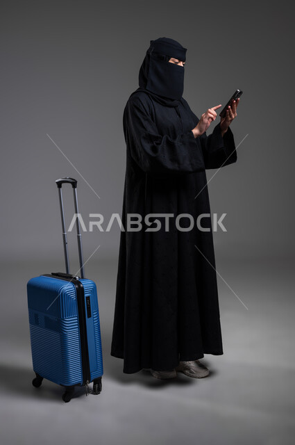 السفر والتعرف إلى البلدان والأماكن الأخرى، الوقوف والاستعداد للرحلات السياحية،  بورتريه لامرأة عربية خليجية سعودية مسلمة منقبة ترتدي العباءة السوداء تمسك الهاتف المحمول وبجانبها حقيبة السفر الزرقاء، الجسم بالطول الكامل، خلفية رمادية