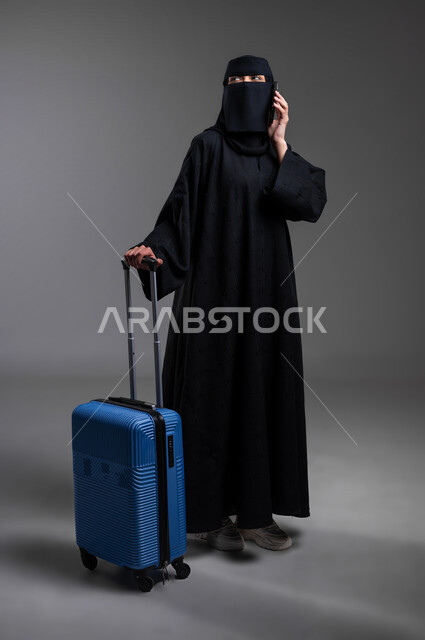 الإمساك بالهاتف المحمول وإجراء مكالمة هاتفية، الوقوف والاستعداد للرحلات السياحية، الذهاب في مغامرات لاستكشاف البلدان والأماكن الأخرى، بورتريه لامرأة عربية خليجية سعودية مسلمة منقبة ترتدي العباءة السوداء تجر حقيبتها السفر، الجسم بالطول الكامل، خلفية رمادية