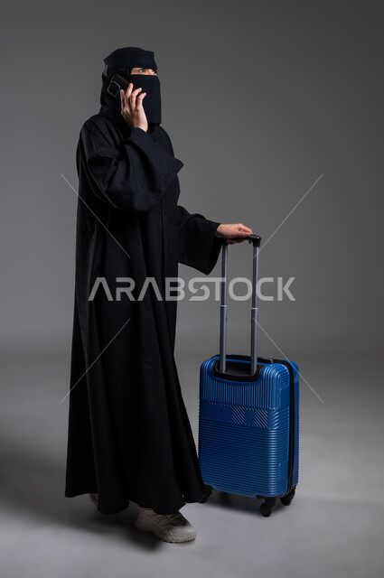 الوقوف والاستعداد للرحلات السياحية، الإمساك بالهاتف المحمول وإجراء مكالمة هاتفية، الذهاب في مغامرات لاستكشاف البلدان والأماكن الأخرى، بورتريه لامرأة عربية خليجية سعودية مسلمة منقبة ترتدي العباءة السوداء تجر حقيبتها السفر، الجسم بالطول الكامل، خلفية رمادية