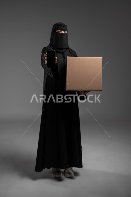 Displaying a blank black screen using a mobile phone, explanatory panels and educational aids, marketing offers and advertisements, a rectangular mockup, a portrait of a veiled Saudi Arabian Gulf Muslim woman wearing an abaya, standing straight and holding her hands, a blank beige canvas, full-length body, gray background