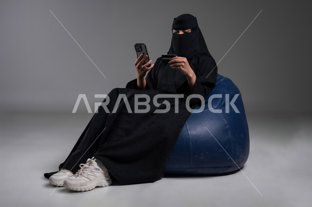 الإمساك بالكرت المصرفي والتأكد من المعلومات الشخصية، استخدام الأجهزة الحديثة والتكنولوجيا والاستفادة منها في المجالات العملية، بورتريه لامرأة عربية خليجية سعودية مسلمة منقبة ترتدي العباءة السوداء تجلس على الكرسي الأزرق المريح بإيماءات الاسترخاء، خلفية رمادية