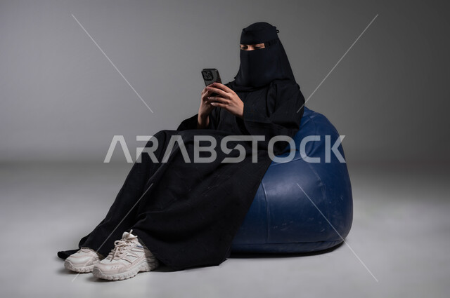 الاستمتاع بقضاء الأوقات، التواصل والدردشة مع الأهل والأصدقاء، استخدام الأجهزة الحديثة والتكنولوجيا في مختلف المجالات العملية، بورتريه لامرأة عربية خليجية سعودية مسلمة منقبة ترتدي العباءة السوداء تجلس على الكرسي الأزرق المريح بإيماءات الاسترخاء، خلفية رمادية