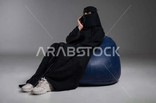 التواصل والدردشة مع الأهل والأصدقاء، إجراء مكالمات هاتفية مهمة، استخدام الأجهزة الحديثة والتكنولوجيا في مختلف المجالات العملية، بورتريه لامرأة عربية خليجية سعودية مسلمة منقبة ترتدي العباءة السوداء تجلس على الكرسي الأزرق المريح بإيماءات الاسترخاء، خلفية رمادية