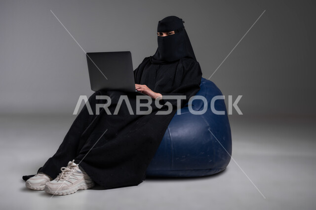 التواصل والدردشة مع الأهل والأصدقاء، استخدام الأجهزة الحديثة والتكنولوجيا في مختلف مجالات العمل، الجلوس بإيماءات الاسترخاء على كرسي أزرق مريح، بورتريه لامرأة عربية خليجية سعودية مسلمة منقبة ترتدي العباءة السوداء تعمل على الحاسوب المحمول، خلفية رمادية