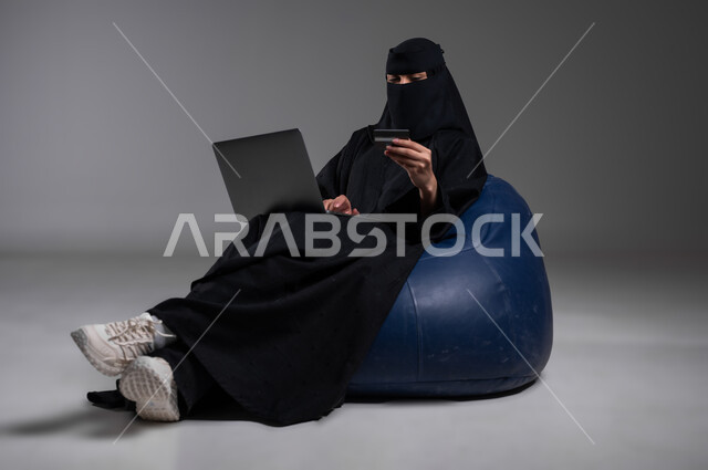 استخدام الأجهزة الحديثة والتكنولوجيا في مختلف مجالات العمل، الجلوس بإيماءات الاسترخاء على كرسي أزرق مريح، بورتريه لامرأة عربية خليجية سعودية مسلمة منقبة ترتدي العباءة السوداء تعمل على الحاسوب المحمول، التواصل والدردشة مع الأهل والأصدقاء، خلفية رمادية