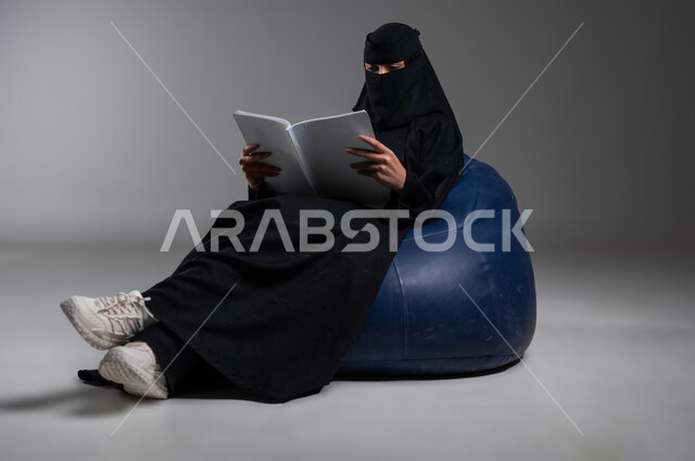 التزود بالثقافة والفكر، قراءة الكتب والروايات العلمية والأدبية في أوقات الفراغ، التعرف على معلومات جديدة قيمة، بورتريه لامرأة عربية خليجية سعودية مسلمة منقبة ترتدي العباءة السوداء تجلس على الكرسي الأزرق المريح بإيماءات الاسترخاء، خلفية رمادية