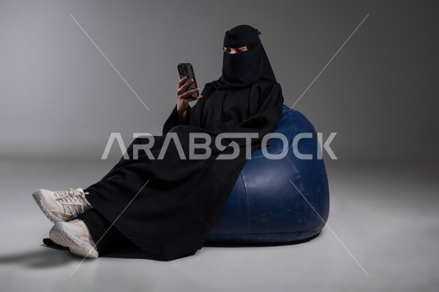 Following the latest developments and news around the world, sitting with relaxing gestures on a comfortable blue chair, portrait of a veiled Saudi Gulf Arab Muslim woman wearing a black abaya making a phone call using a mobile phone, communicating with family and friends, gray background