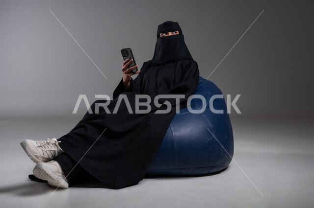 Taking advantage of modern technologies, sitting with relaxing gestures on a comfortable blue chair, portrait of a veiled Saudi Gulf Arab Muslim woman wearing a black abaya, holding a mobile phone in her hand, communicating with family and friends, following the latest developments and news around the world, gray background