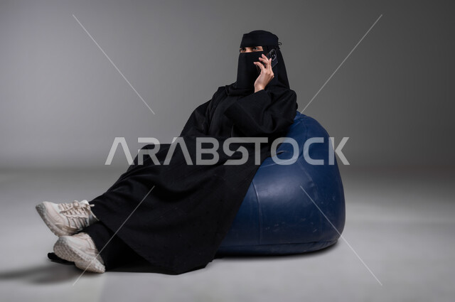 Sitting with relaxing gestures on a comfortable blue chair, portrait of a veiled Saudi Gulf Arab Muslim woman wearing a black abaya making a phone call using a mobile phone, communicating with family and friends, following the latest developments and news around the world, gray background
