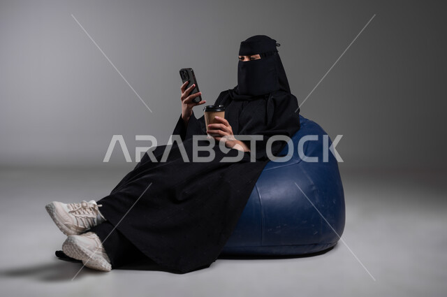 Getting a cup of hot coffee, spending enjoyable times, sitting with relaxing gestures on a comfortable blue chair, portrait of a veiled Saudi Gulf Arab Muslim woman wearing a black abaya, holding a mobile phone in her hand, communicating with family and friends, gray background