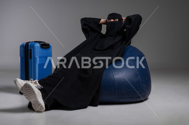 الاستعداد للذهاب في رحلات ممتعة، الجلوس بإيماءات الاسترخاء بانتظار وقت الرحلة، بورتريه لامرأة عربية خليجية سعودية مسلمة منقبة ترتدي العباءة السوداء تجلس على كرسي مريح وبجانبها حقيبة سفر زرقاء، مفهوم السياحة والسفر والاستكشاف، خلفية رمادية