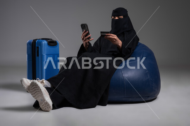 التأكد من وقت الرحلة والمعلومات الهامة، الاستعداد للذهاب في رحلات ممتعة، استخدام الهاتف المحمول في وقت الانتظار، بورتريه لامرأة عربية خليجية سعودية مسلمة منقبة ترتدي العباءة السوداء تجلس على كرسي مريح وبجانبها حقيبة سفر زرقاء، خلفية رمادية
