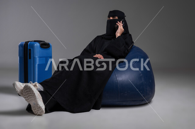 إجراء مكالمة هاتفية والدردشة مع الأصدقاء، الاستعداد للذهاب في رحلات ممتعة، استخدام الهاتف المحمول خلال وقت الانتظار، بورتريه لامرأة عربية خليجية سعودية مسلمة منقبة ترتدي العباءة السوداء تجلس على كرسي مريح وبجانبها حقيبة سفر زرقاء، خلفية رمادية